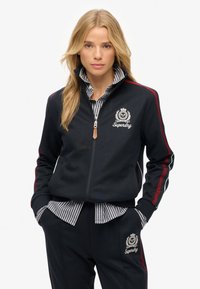 Chaqueta navy con cremallera y franjas laterales rojas y verdes, que presenta un logo en el pecho y un cuello de punto texturizado sobre una camiseta a rayas.
