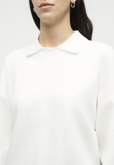 Femme portant un haut blanc uni à manches longues avec un petit col et des boucles d'oreilles en argent en forme d'anneau, montrée des épaules aux lèvres.