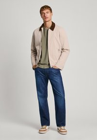 Chaqueta bomber de color beige claro con cuello marrón, camisa verde, vaqueros azules y zapatillas marrones y blancas. El modelo se encuentra de pie con las manos en los bolsillos.