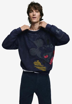 Young man adjusting collar of oversized navy sweatshirt with abstract multicolored graphic, wearing dark pants against plain background.