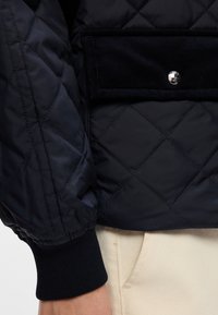 Gepolstete schwarze Pufferjacke mit einer Cord-Tasche und silbernem Druckknopf. Verfügt über elastische Bündchen und eine glatte, glänzende Textur.