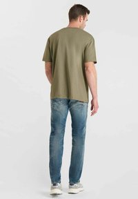 Hombre de pie de espaldas, con una camiseta verde oliva, jeans azules y zapatillas grises, sobre un fondo claro y sencillo.