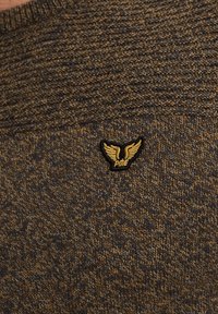 Pull en tricot marron foncé avec un motif texturé. Présente un petit patch noir avec un logo ailé doré sur la zone gauche de la poitrine.