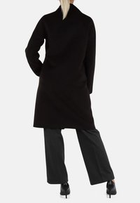 Cappotto nero con silhouette dritta, maniche lunghe e colletto alto. Abbinato a pantaloni grigi a gamba larga e tacchi neri. Texture liscia.