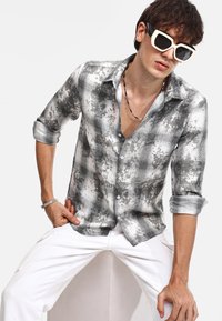 Camicia a quadri grigia e bianca con vestibilità slim, chiusura a bottoni, maniche arrotolate e texture morbida. Indossata con pantaloni bianchi e occhiali da sole grandi.
