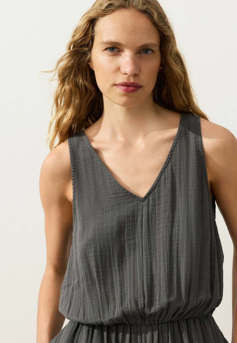Jeune femme aux longs cheveux blonds ondulés porte une robe sans manches gris foncé texturée avec un col en V et une taille élastique.