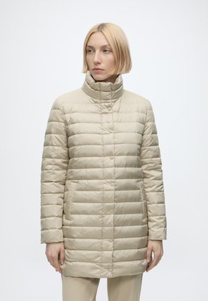 Mujer con cabello rubio corto lleva una chaqueta acolchada beige con cuello alto, de pie frente a un fondo liso y claro.
