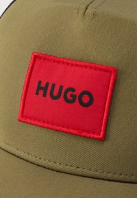 Καπέλο ελιάς πράσινο με κόκκινο ορθογώνιο μπαλώμα που φέρει το μαύρο κείμενο «HUGO». Το ύφασμα έχει υφή με ορατές ραφές κατά μήκος του γείσου.