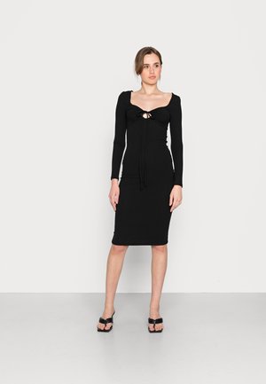 Vestido justo preto de manga longa com um detalhe em forma de buraco no peito, apresentando uma silhueta ajustada e textura suave. Usado com sandálias pretas.