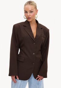 Femme blonde portant un blazer oversize marron foncé avec des boutons et un jean bleu clair, posant devant un fond uni.