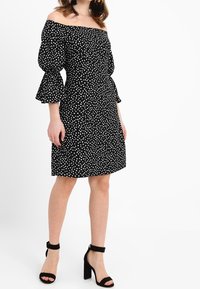 Svart klänning utan axelband med vita prickar, volangärmar, figurnära midja och utsvängd kjol. Matchas med svarta klacksandaler med blockklack.