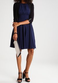 Mujer con un vestido azul marino hasta la rodilla y un cárdigan negro cropped, sosteniendo un clutch beige y usando sandalias de tacón alto negras.