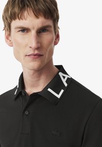 Svart polotröja i mjukt tyg, med en kontrasterande vit stickad krage som har "LA"-bokstäver och en liten svart logotyp på bröstet.