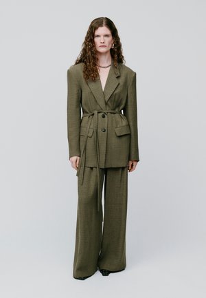 Mujer con cabello largo y rizado lleva un traje de cuadros color verde oliva sobredimensionado con cinturón, pantalones de pierna ancha y zapatos negros, de pie contra un fondo liso.