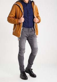 Hombre con una chaqueta con capucha de color mostaza, camisa de botones azul marino, jeans gris desgastados ajustados y zapatos de vestir negros, de pie sobre un fondo blanco.