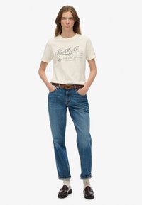Ung kvinde med langt hår iført en cremefarvet T-shirt med grafik, blå jeans med et brunt bælte, hvide sokker og burgunderfarvede loafers, hænderne i lommerne.