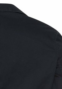 Nærbillede af en sort stofskjorte bagfra, der viser skuldersøm og en del af kraven mod en hvid baggrund.