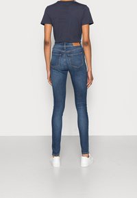 Pantalones ajustados de mezclilla azul oscuro con cintura alta, que cuentan con dos bolsillos traseros y una etiqueta de piel. Combinados con una camiseta básica oscura y zapatillas blancas.