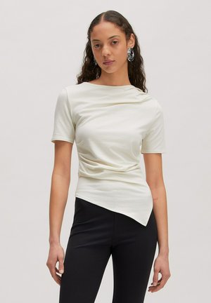 Vrouw met lang krullend haar die een witte top met korte mouwen en een asymmetrische zoom draagt en zwarte broek, staande tegen een effen achtergrond.