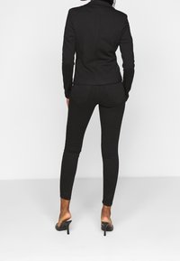 Blazer noir cintré et jeans noirs slim, en tissu lisse avec des détails de couture minimalistes. Le mannequin porte des talons ouverts noirs.
