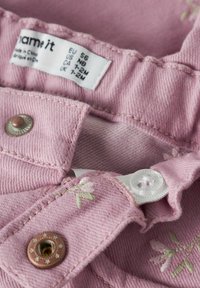 Rožinės spalvos džinsinis audinys su balta ir bronzos spalvos furnitūra, subtiliu gėlių siuvinėjimu ir matoma priežiūros etiketė su dydžio informacija.