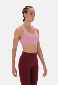 Růžová sportovní podprsenka s vlnitým lemem, výstřihem do U a přiléhavým střihem, kombinovaná s vysoce pasovanými burgundskými legínami. Hladký materiál, minimální detaily.