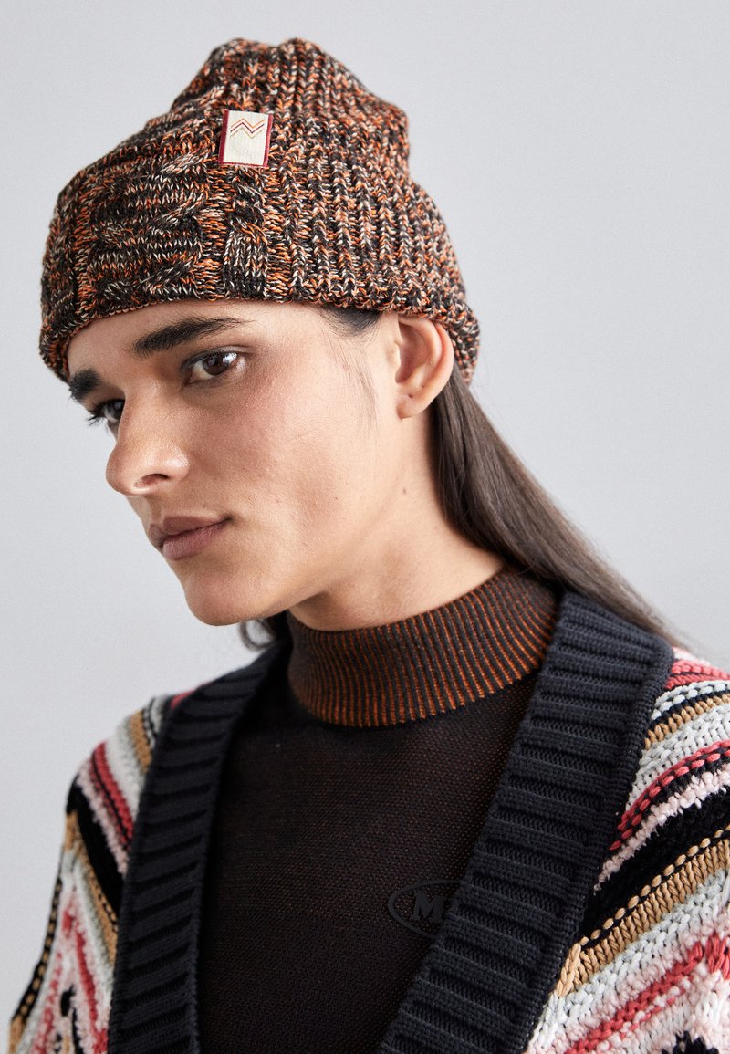 Knit beanie in orange, brown, and black with a textured pattern and a small brand tag. Worn with a dark, ribbed turtleneck and multi-colored cardigan.