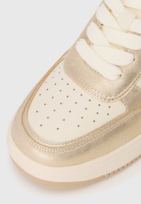 Front section of beige and gold sneaker with perforated toe box, thick white laces, and textured sole visible against white background.