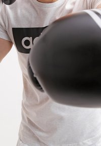 Guantes de boxeo negros con franjas blancas, usados por una persona con una camiseta gris claro que presenta un logo negro, contra un fondo liso.
