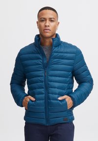 Blau gepolsterte Jacke mit hohem Kragen, Reißverschluss und horizontalen Steppnähten. Verfügt über Seitentaschen und eine glatte Textur.