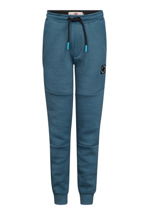Junge steht, trägt ein blaues Kurzarmhemd und passende Hosen mit weißen Sneakers vor einem schlichten hellen Hintergrund.