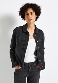 Mustahi denim takki, jossa on klassinen kaulus, nappilukitus edessä ja kaksi rintataskua. Päällä valkoinen hihaton paita ja vastaavat mustat farkut.