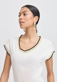 Top bianco a maniche corte con scollatura a V, rifinito con bordi ondulati dorati e neri. Il modello indossa orecchini pendenti con accenti rosa e verdi.