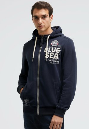 Junger Mann trägt eine dunkelblaue Kapuzenjacke mit Reißverschluss, weißen Kordeln und bedrucktem Text auf Brust und Tasche, steht mit den Händen in den Taschen.