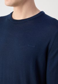 Tengerészkék kötött pulóver kerek nyakkivágással és diszkrét, azonos színben hímzett GANT logóval a mellrészén. Sima textúra.