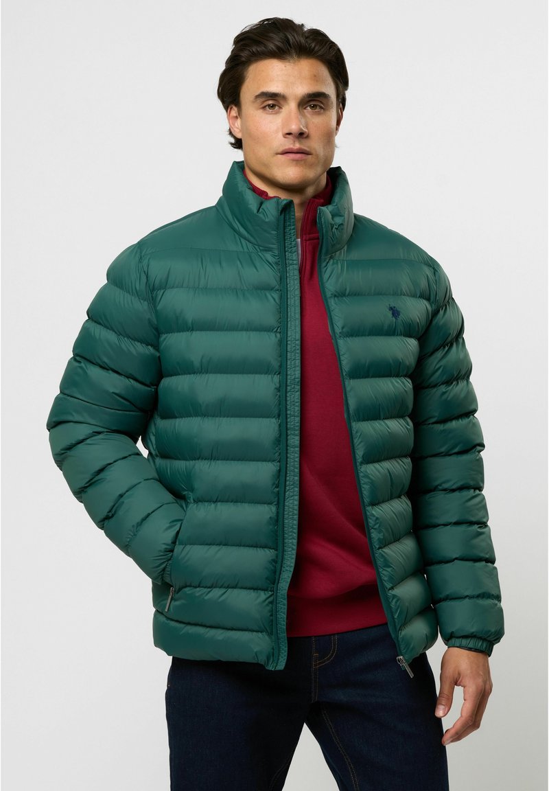 Gevoerde groene jas met een hoge kraag, voorzien van een ritssluiting aan de voorkant en elastische manchet, gedragen over een kastanjebruine trui, gepresenteerd op een model.