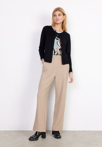 Black cardigan with button closure, paired with beige high-waisted trousers and black platform shoes. Patterned top visible underneath.