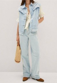 Hellblauer Denim-Weste mit weitem Schnitt, weiße Bluse und hellblaue Jeans. Geflochtene Tasche in der rechten Hand. Braune Stiefeletten mit Absatz.