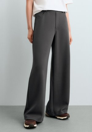 Personne portant un pantalon large gris foncé et des baskets marron, debout contre un mur et un sol gris clair.