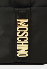 Czarna torba z nylonu z wyraźnym złotym akcentem w postaci logo "Moschino". Design charakteryzuje się gładką teksturą i solidnymi elementami metalowymi.