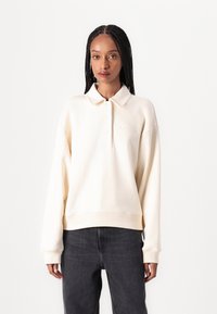Krämfärgad, oversized polo-sweatshirt med en knappslå och ribbade muddar. Har en subtil broderad logo på bröstet.