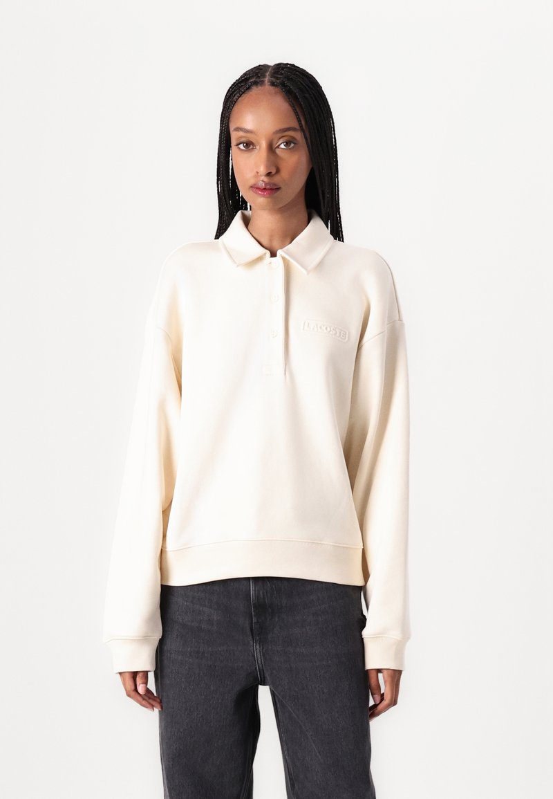 Krämfärgad, oversized polo-sweatshirt med en knappslå och ribbade muddar. Har en subtil broderad logo på bröstet.