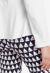 Witte lange mouwen top boven zwarte leggings met een repetitief wit zeilbootpatroon, dat contrast en textuur benadrukt.