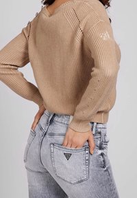 Beige knitted sweater with ribbed texture, featuring shoulder button accents. Grey distressed jeans with a logo patch on the back pocket.
