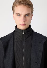 Veste noire matelassée avec un col haut, équipée d'une fermeture éclair à l'avant. Portée sous un manteau texturé gris foncé.