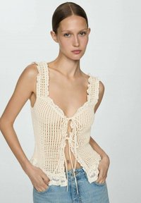 Top in crochet crema con ampie spalline, scollatura a V profonda, chiusura frontale con laccetti e bordo in pizzo ondulato all'orlo. Abbinato a jeans in denim blu.