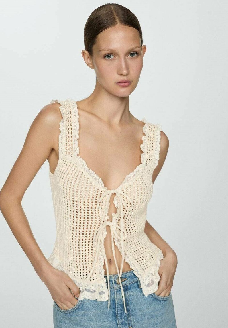 Top in crochet crema con ampie spalline, scollatura a V profonda, chiusura frontale con laccetti e bordo in pizzo ondulato all'orlo. Abbinato a jeans in denim blu.