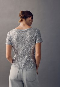 Top a maniche corte con paillettes argentate e vestibilità ampia, con scollo arrotondato e schiena trasparente, abbinato a pantaloni in denim grigio chiaro.