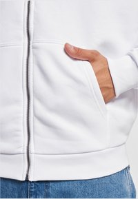 White zip-up sweatshirt with a smooth texture, side pockets, and ribbed cuffs. Features a silver zipper and a simple, clean design.
