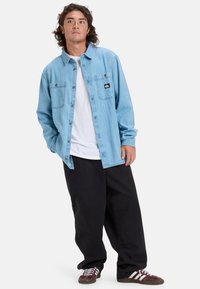 Lichtblauw denim overhemden met knopen en twee borstzakken, gedragen over een wit T-shirt, in combinatie met zwarte losse broek en bruine sneakers.